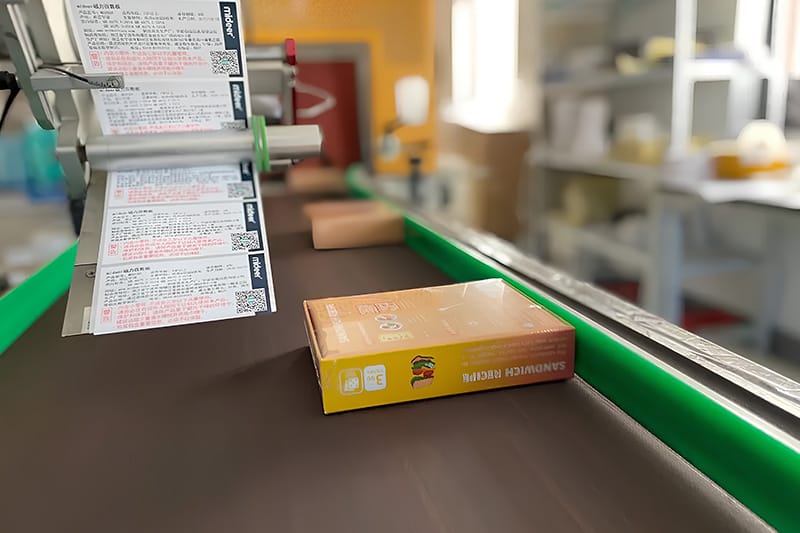 Automated labeling machine applying labels to sandwich recipe boxes on conveyor belt.